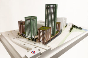 Architectural sales model 1:150 scale. Five highrise buildings painted green and brown brick. White offiste buildings. a tube station is in the foreground. Architectural model is animated with trees.