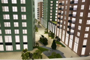 Close up image of an Architectural sales model, 1:150 scale. View down a raod inbetween two highrise buildings, painted green and brown brick. Roads are alser etched and animated wiht etched vellum trees.